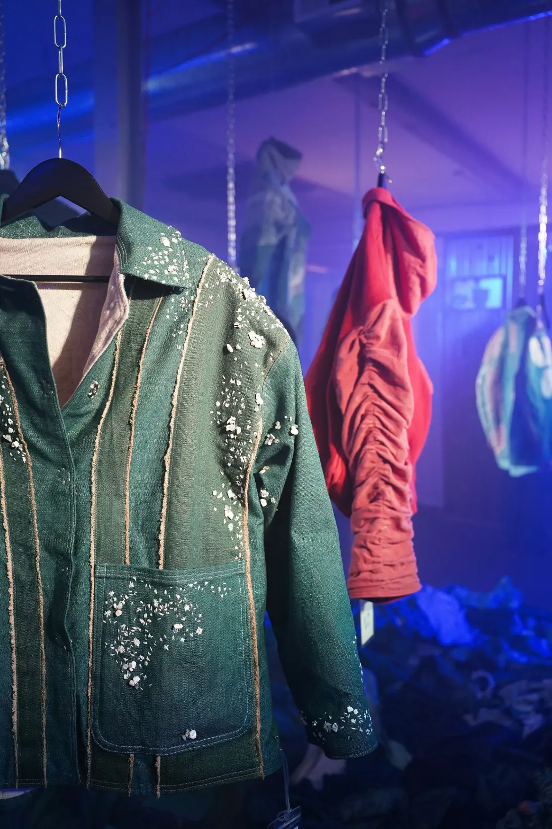 An embellished green jacket with other garments in the exhibition behind it